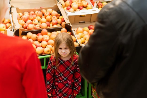 little girl looks angrily towards mom and dad while standing beside a supermarket food stall