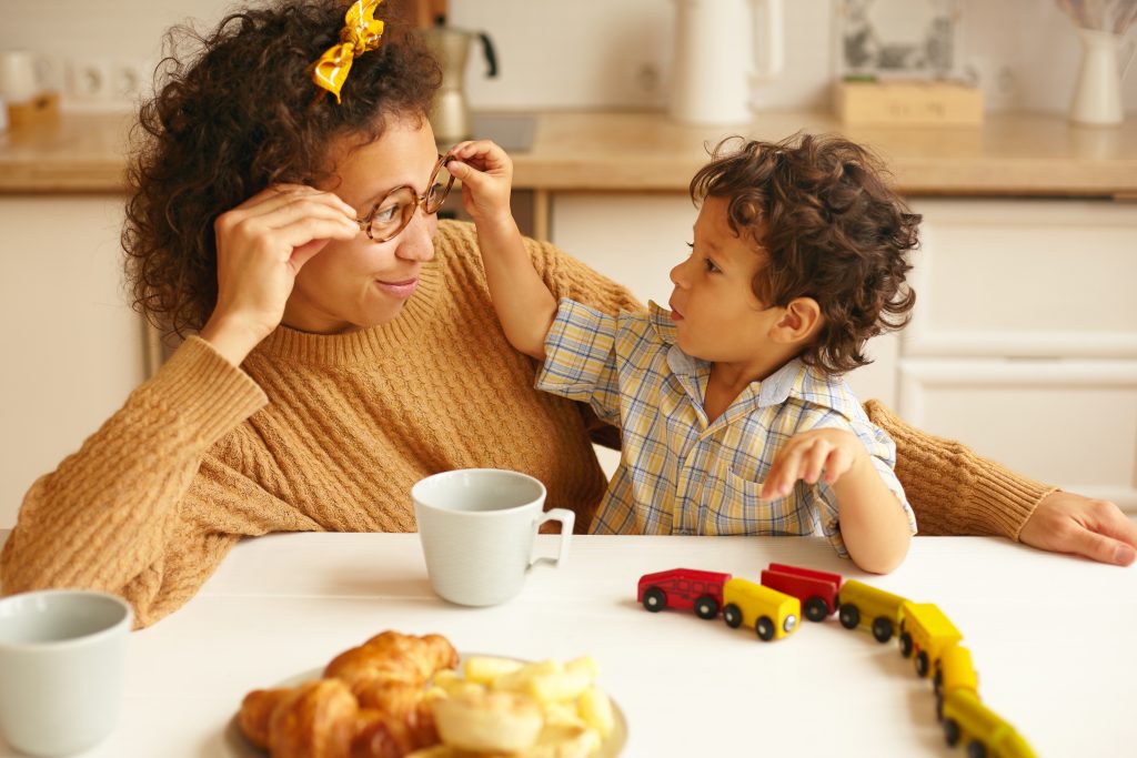 Children, kids, happy childhood, family bonds and parenting concept. Picture of attractive young Hispanic woman having coffee at ktichen table and smiling while infant son taking off her eyeglasses