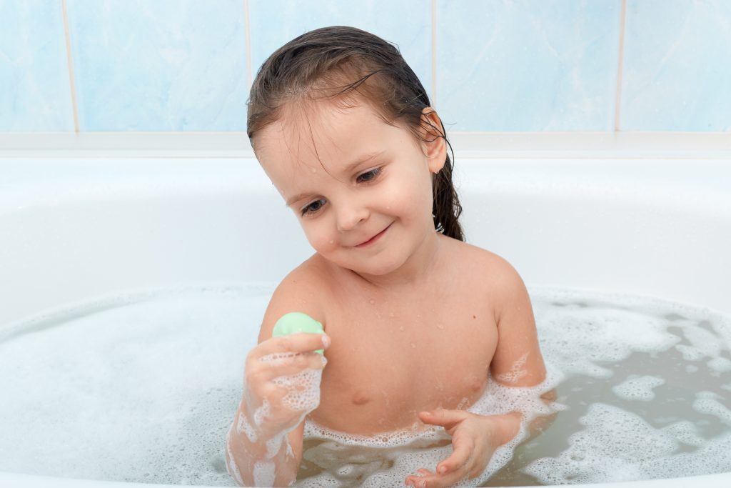 Happy baby taking bath alone, playing with foam bubbles and her new toy. Little child having fun in bathtub. Infant washing and bathing. Hygiene and care for young children before going to bed.