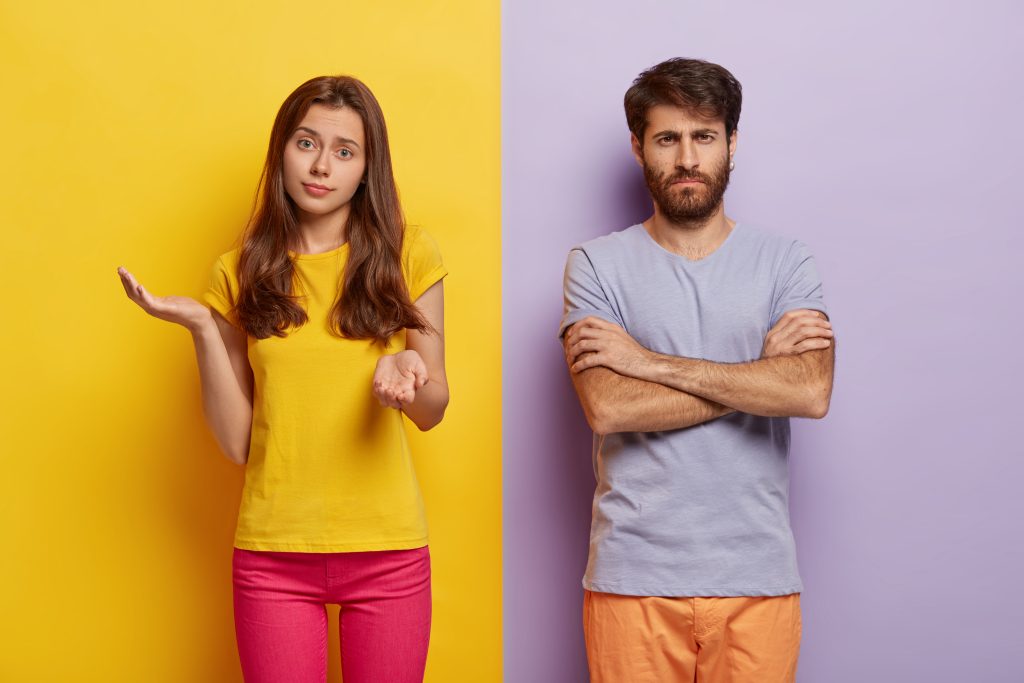 Hesitant young woman spreads hands with doubt, wears casual clothes, dissatisfied man keeps hands crossed, discontent with something, stands against two colored studio wall. Negative emotions