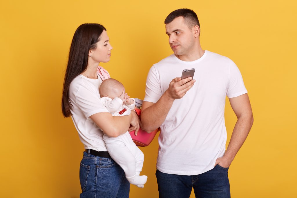 Mother father and baby standing against yellow background, adorable female looking at husband, making complaint, male with phone in hands checking social networks, does not help with newborn daughter.