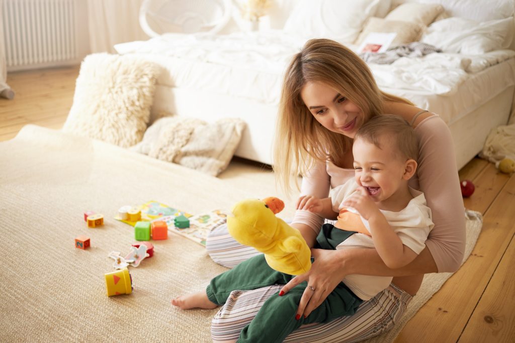 Pretty sister spending time with her baby brother, sitting on floor in bedroom. Beautiful young babysitter playing with little boy indoors, holding stuffed toy duck. Infancy, childcare and motherhood